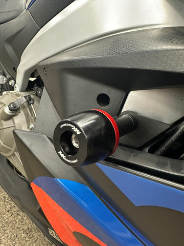 A close-up of a motorcycle side fairing accessory with a black cylindrical handlebar bar end featuring a red accent ring and white branding text, mounted against a silver and blue motorcycle body panel.
